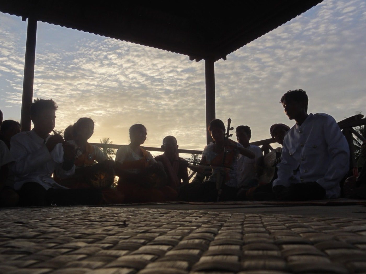 Musicians with traditional instruments seated in a circle under a pavilion roof at dusk.