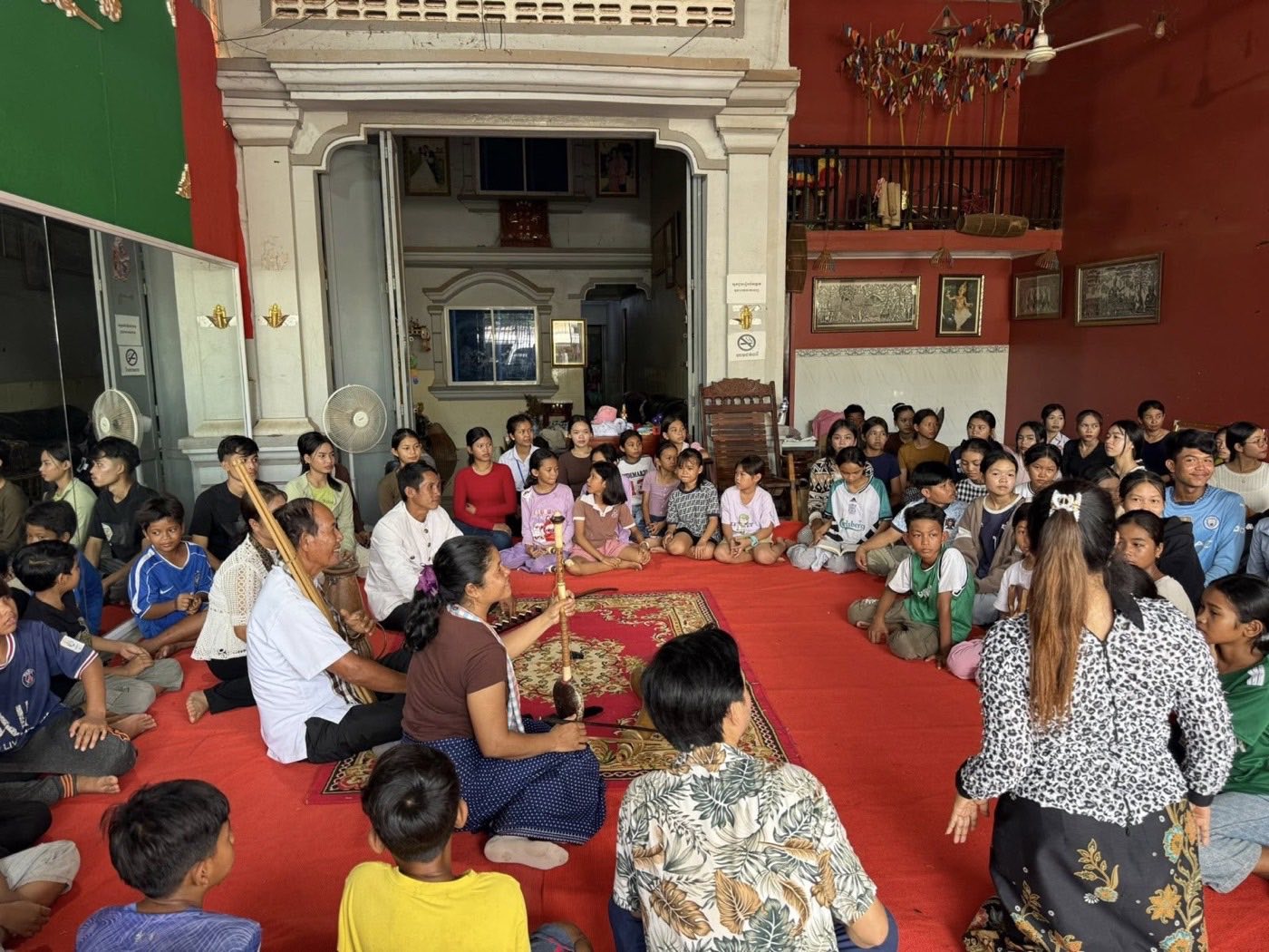 A packed hall at the Samut School of Music and Dance: children seated on red carpet around musicians.